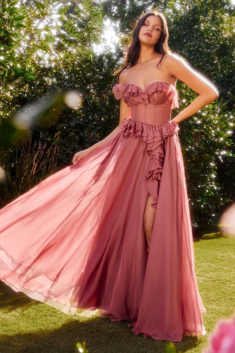 A woman stands outdoors in sunlight, surrounded by lush greenery, wearing the Andrea and Leo A1341 Strapless Ruffled Prom Dress with a high slit and flowing pink fabric.